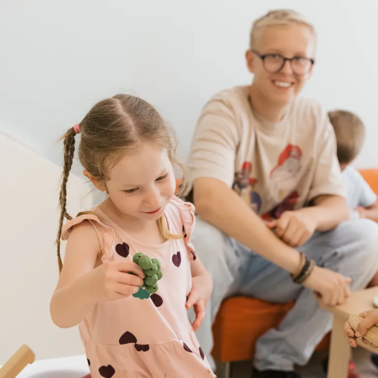 Ein Kind spielt in der Kinderkrippe mit einer kleinen Spielfigur, während eine Betreuungsperson im Hintergrund sitzt.