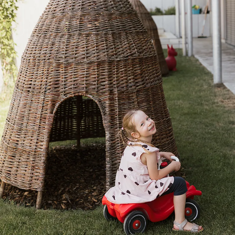 Ein Kind sitzt im Garten der Kinderkrippe auf einem roten Rutschauto vor einem großen Spielhaus aus Weidengeflecht.